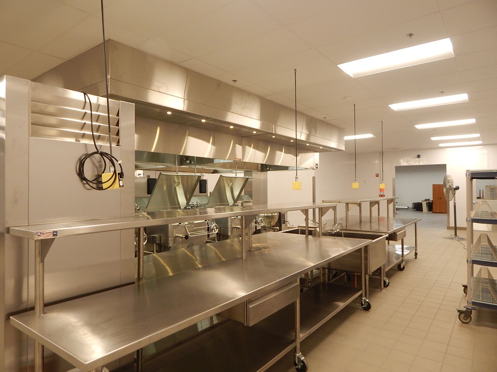 A commercial kitchen featuring long stainless steel prep tables, overhead ventilation hoods, and tiled floors.