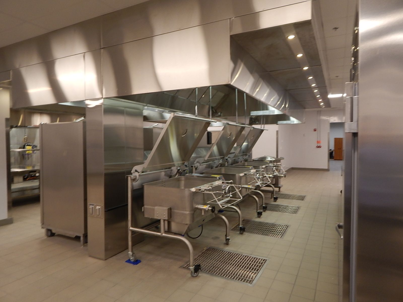 A commercial kitchen features a row of stainless steel tilting skillets beneath large exhaust hoods.