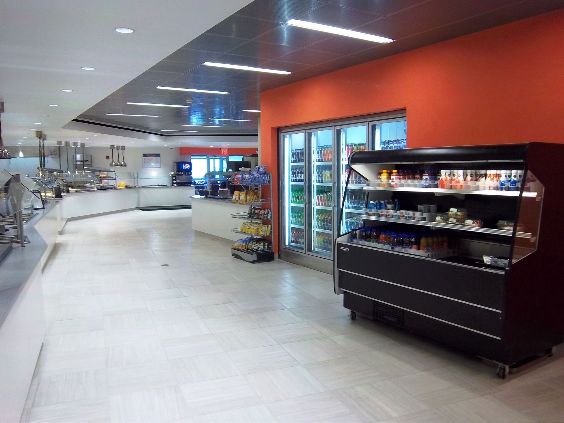 A cafeteria interior featuring white floors, a long serving counter, a bright orange wall, and refrigerated drink displays.