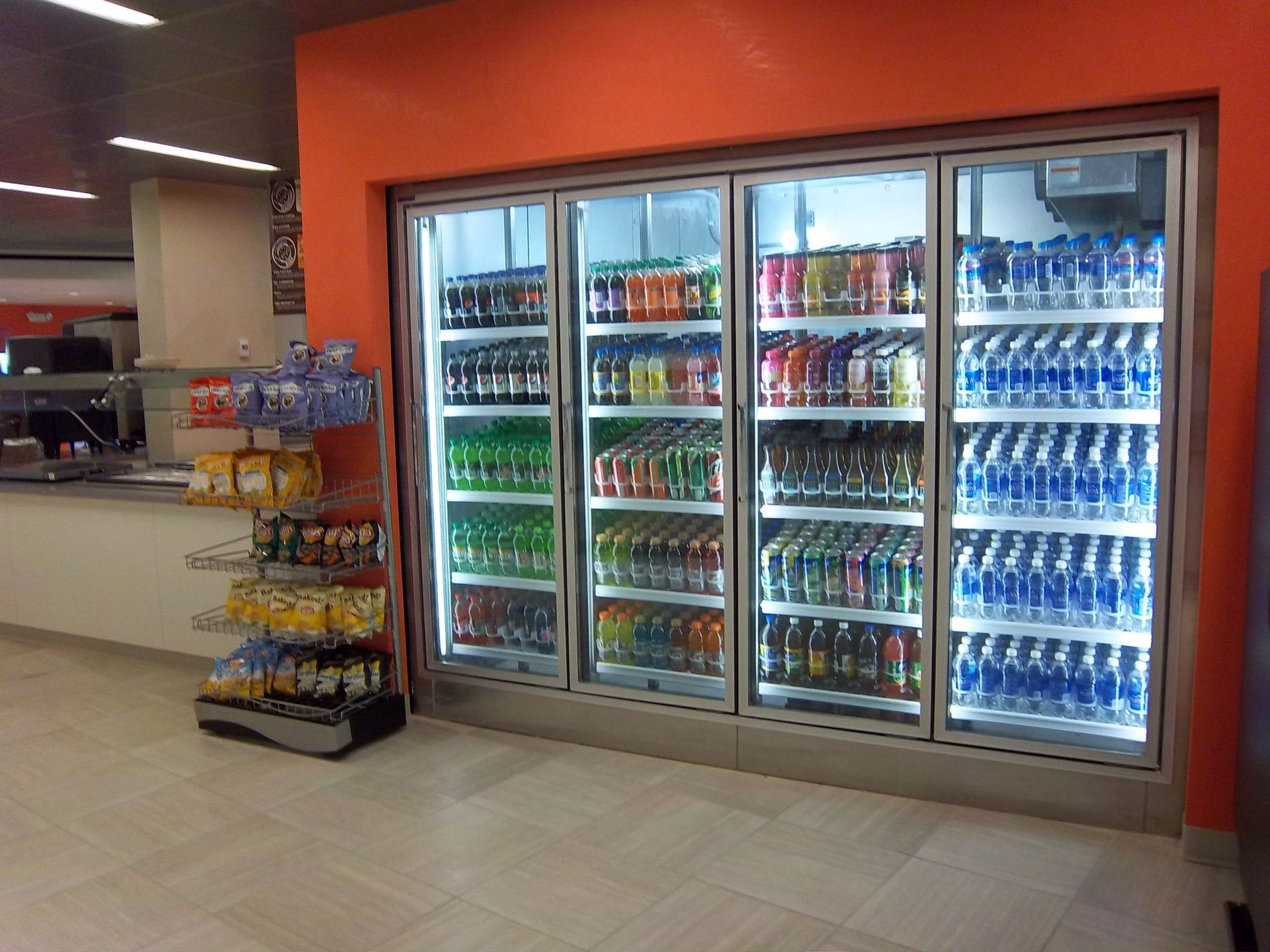 A large refrigerated display case filled with rows of bottled drinks next to a snack rack against an orange wall.
