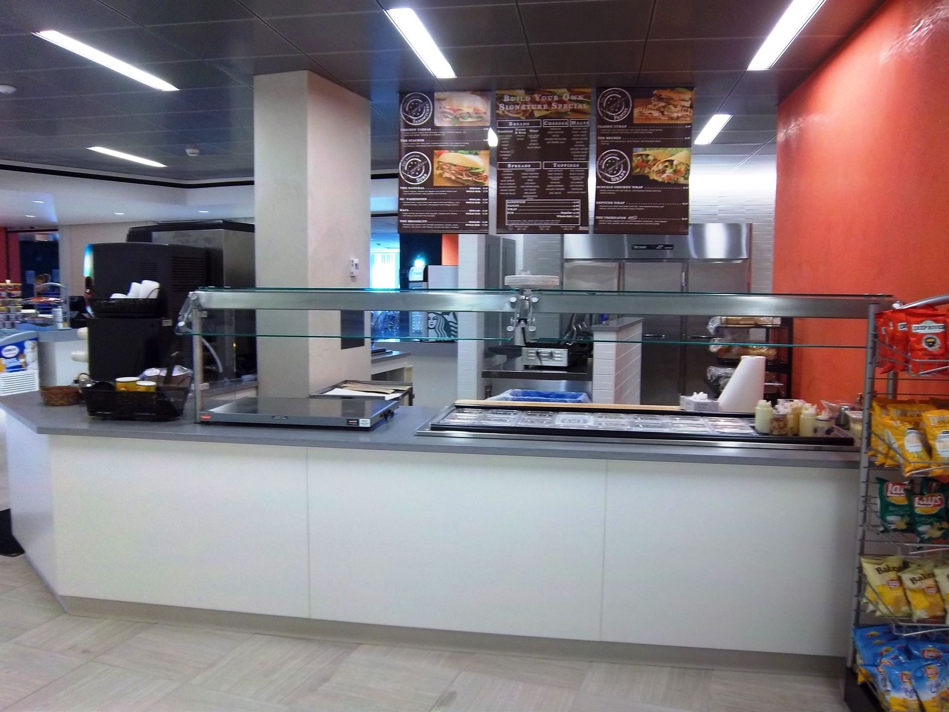 A service counter at a cafe with a white base, glass sneeze guard, menu board above, and a snack rack on the right.