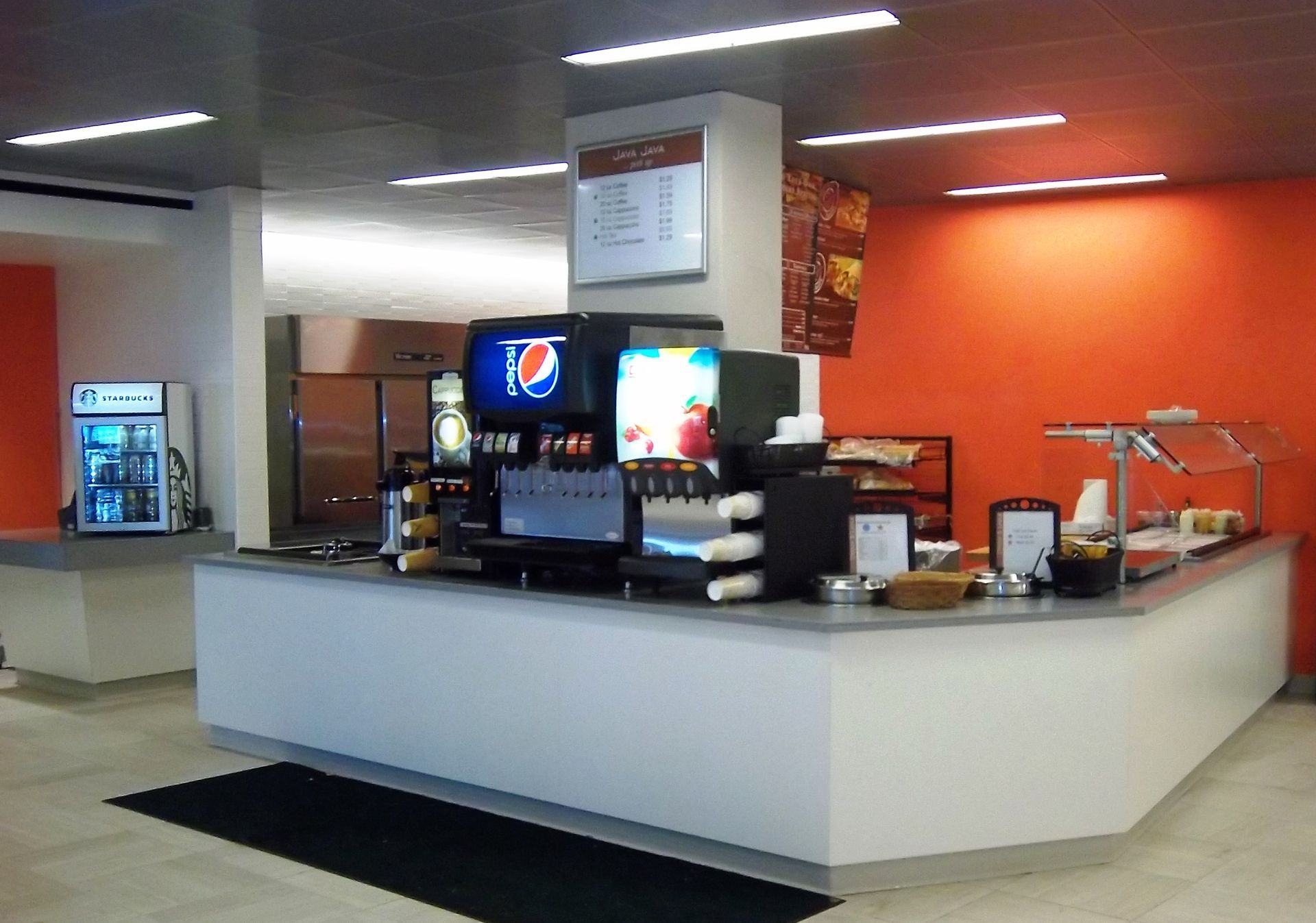 A food service station with a white counter, Pepsi fountain drinks, and an orange accent wall in a bright, modern room.