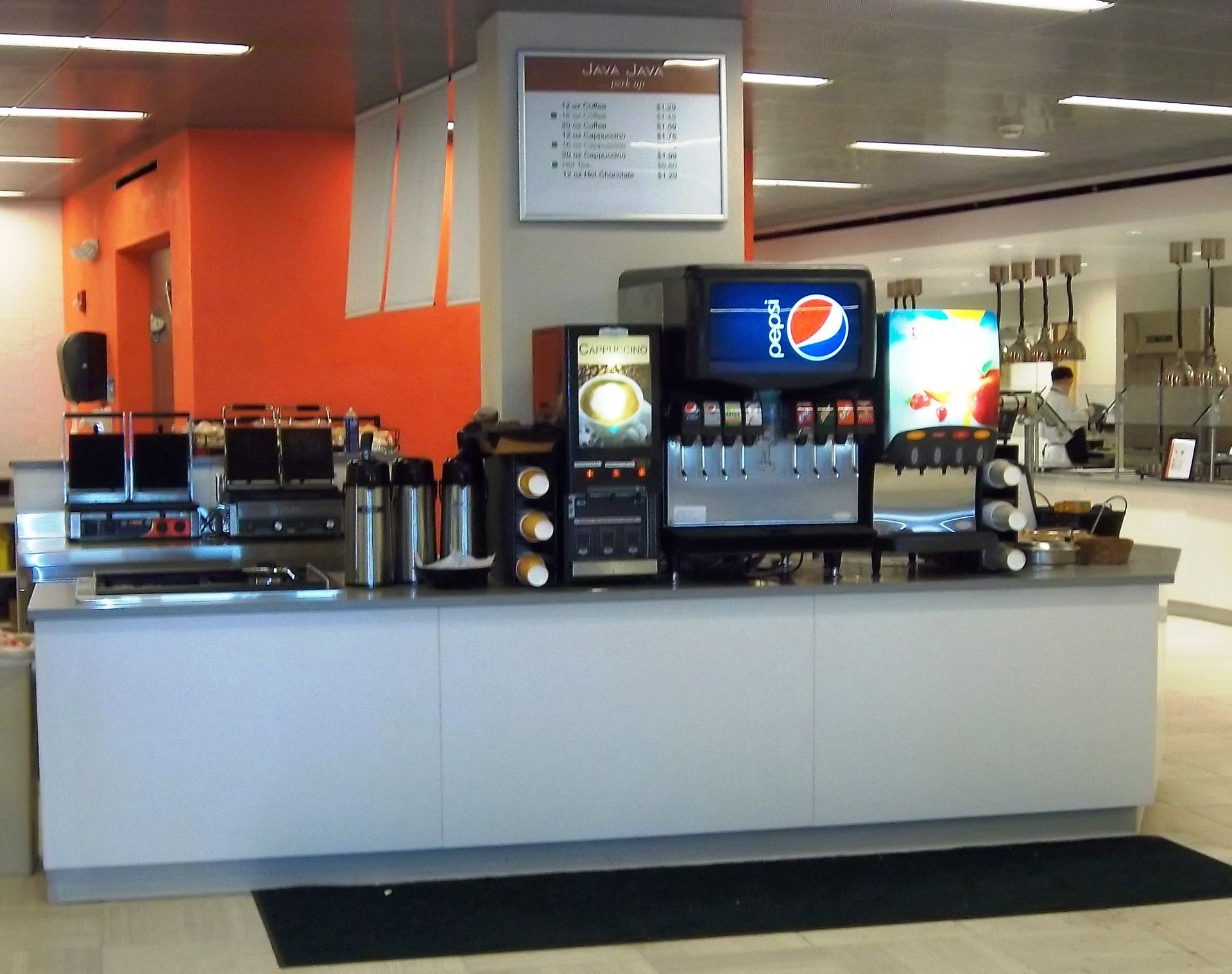 A cafeteria drink station with a soda fountain, coffee machine, and waffle irons against an orange wall.