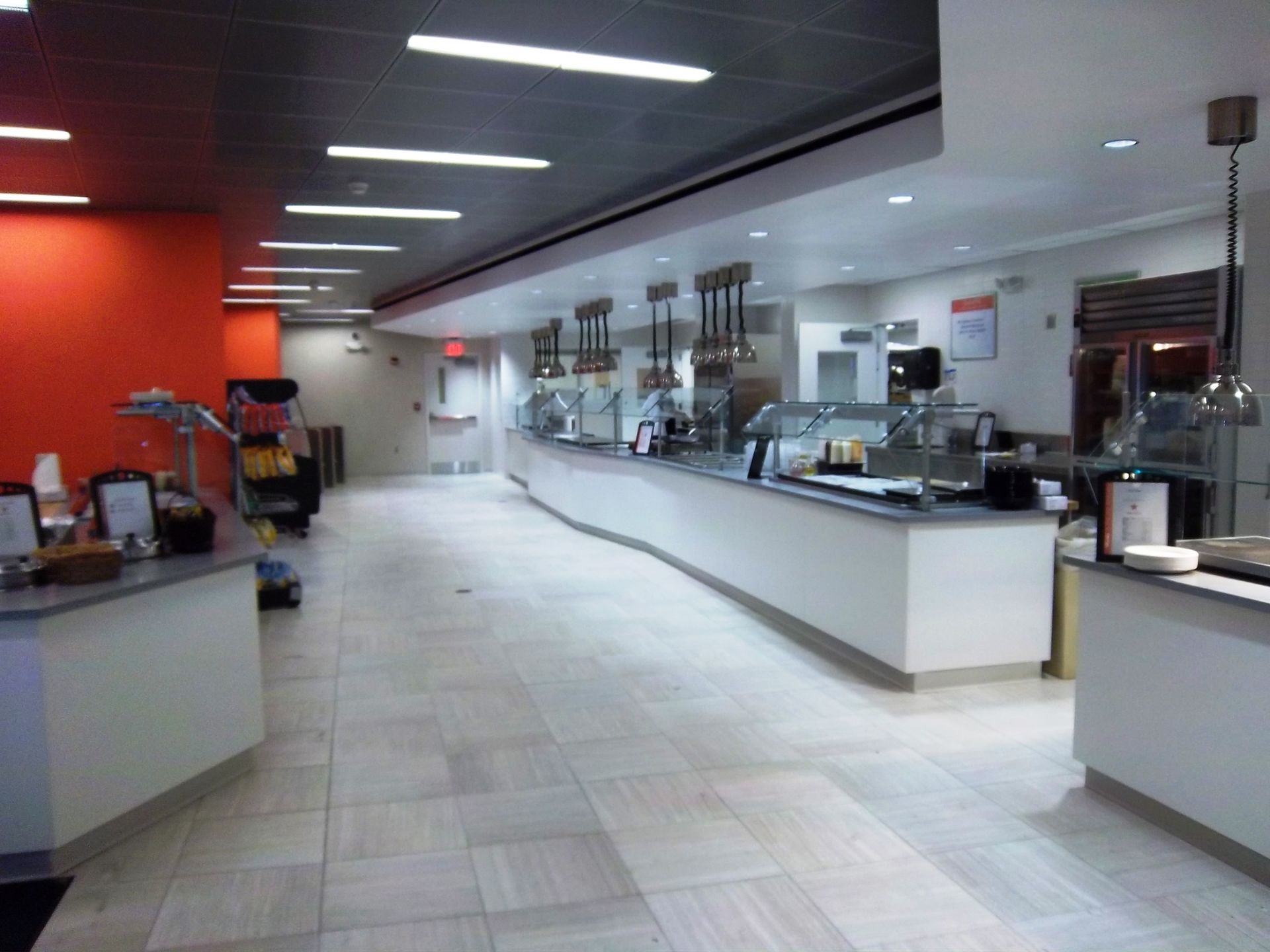 A clean, modern cafeteria with a long white service counter, tiled floor, and recessed lighting against an orange wall.