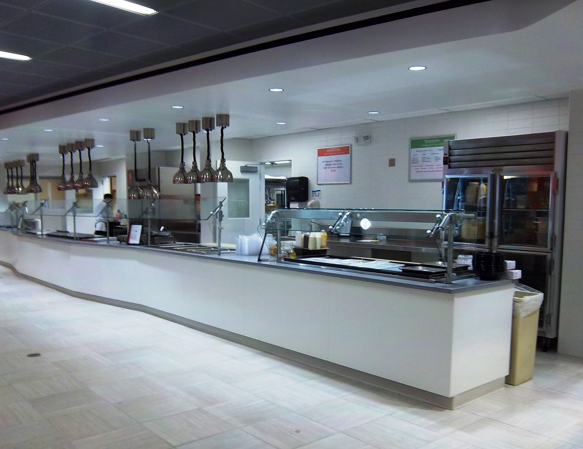 A white, curved food service counter with metal heat lamps and a stainless steel refrigerator in a brightly lit cafeteria.