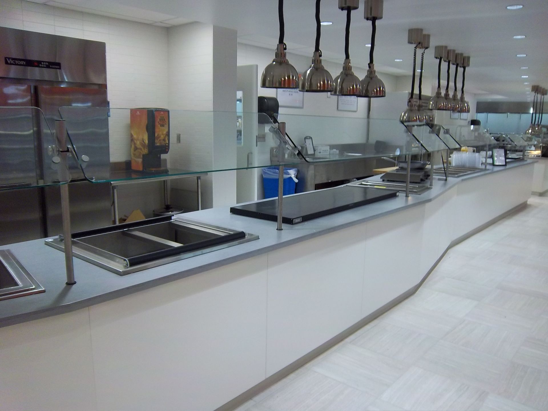 A clean, modern cafeteria serving counter with white cabinets, gray countertops, and overhead heat lamps.