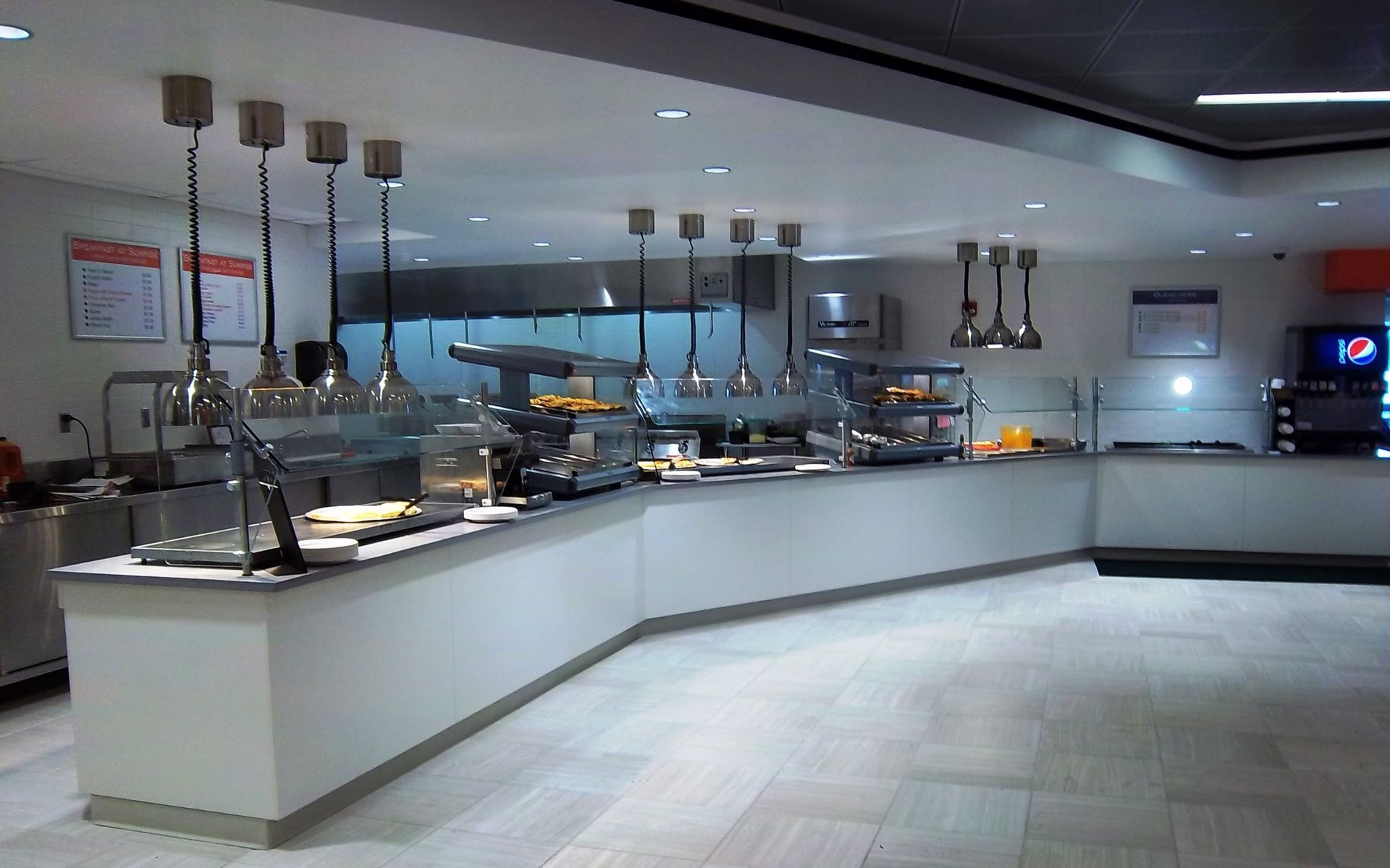 An empty cafeteria buffet counter featuring white serving stations, multiple hanging lights, and a tiled floor.
