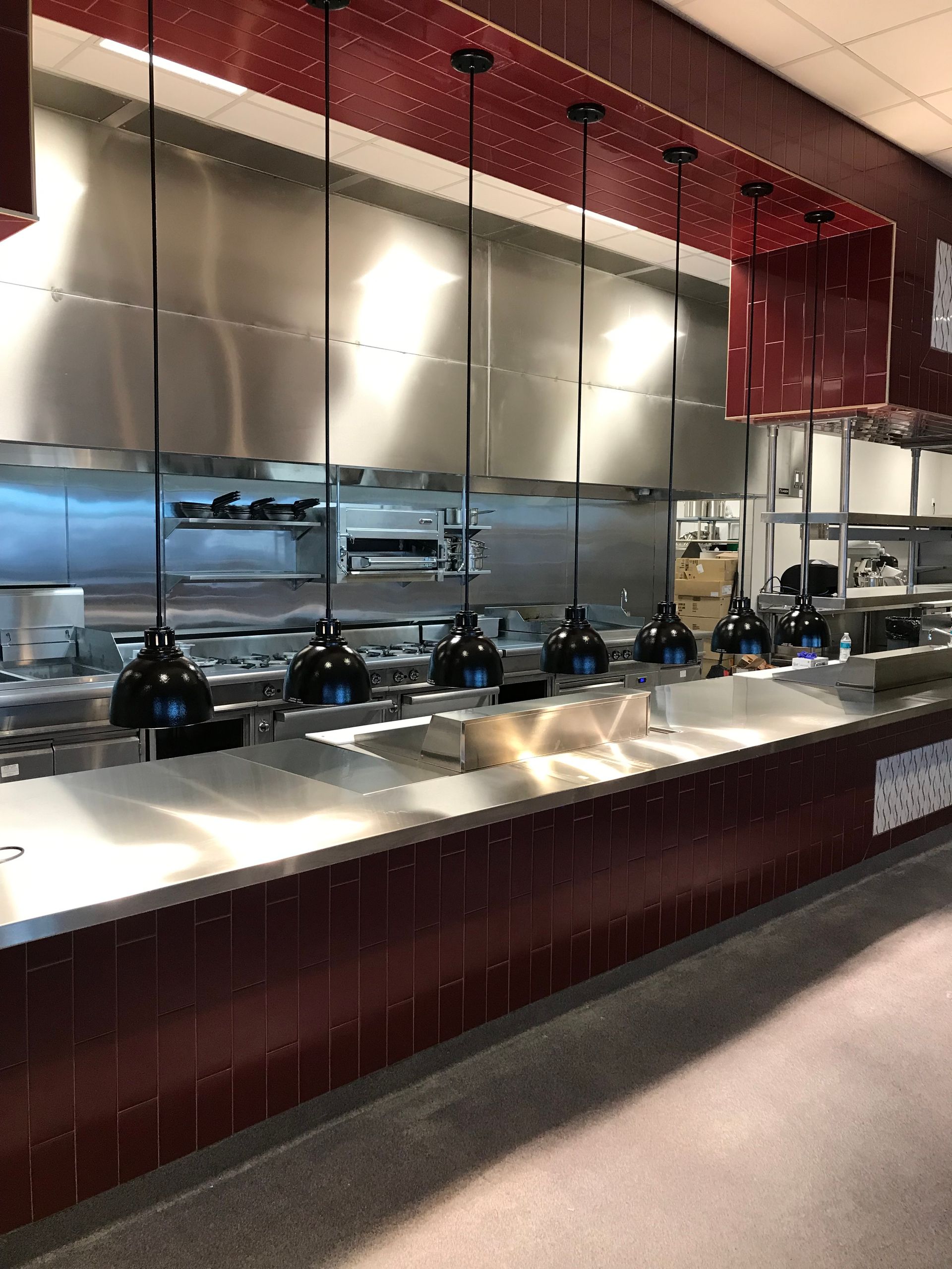 A commercial kitchen line with a metallic counter, stainless steel backsplash, and a row of black pendant heat lamps.