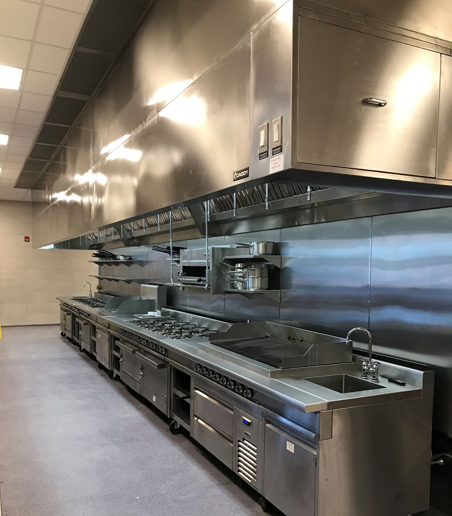 A commercial kitchen featuring stainless steel prep surfaces, cooking ranges, and a large overhead ventilation hood.