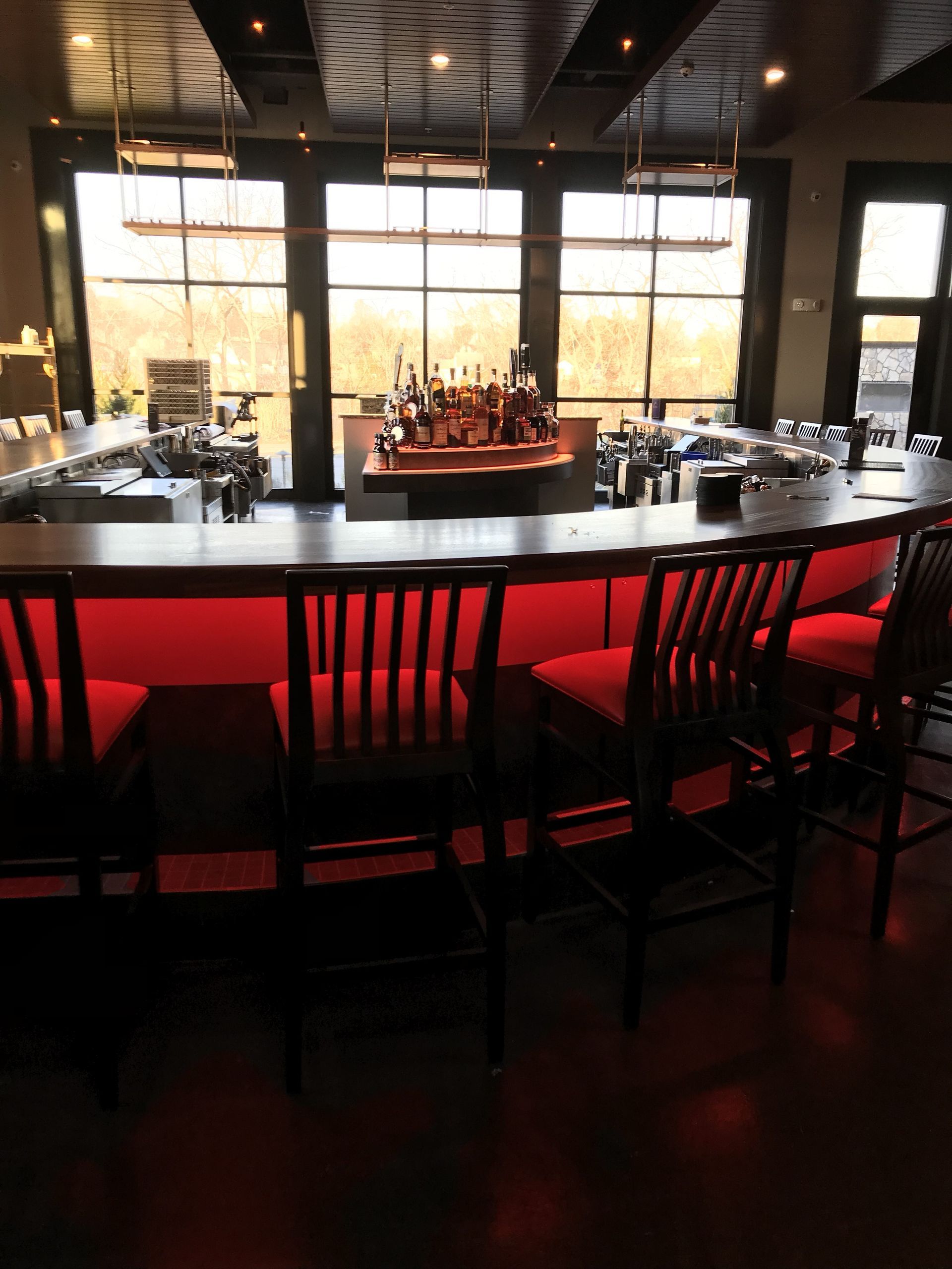 A curved bar with red under-lighting and matching bar stools faces large windows in a dimly lit room.