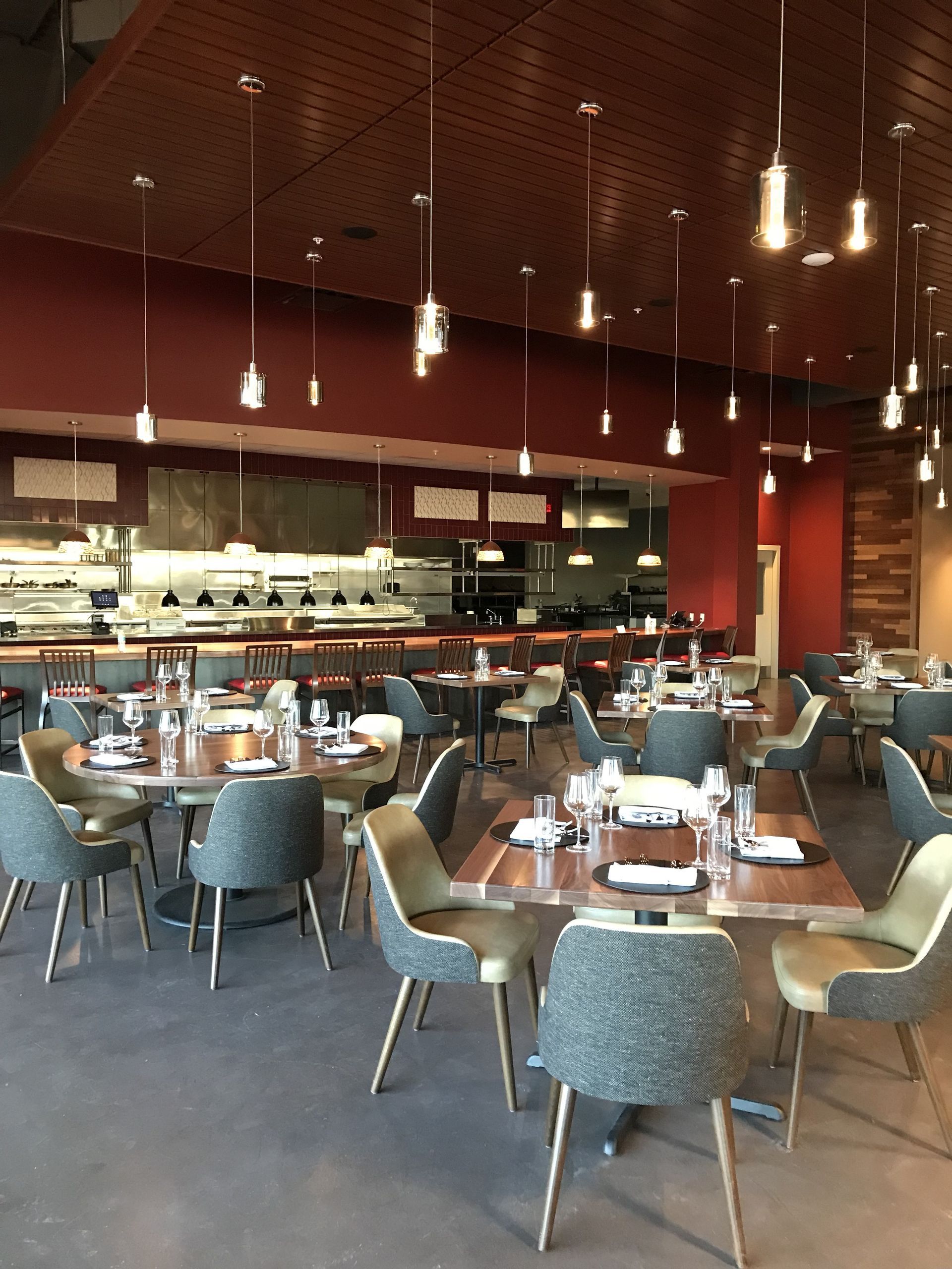 Modern restaurant dining room with dark red walls, wood ceiling, pendant lights, and round tables with gray chairs.
