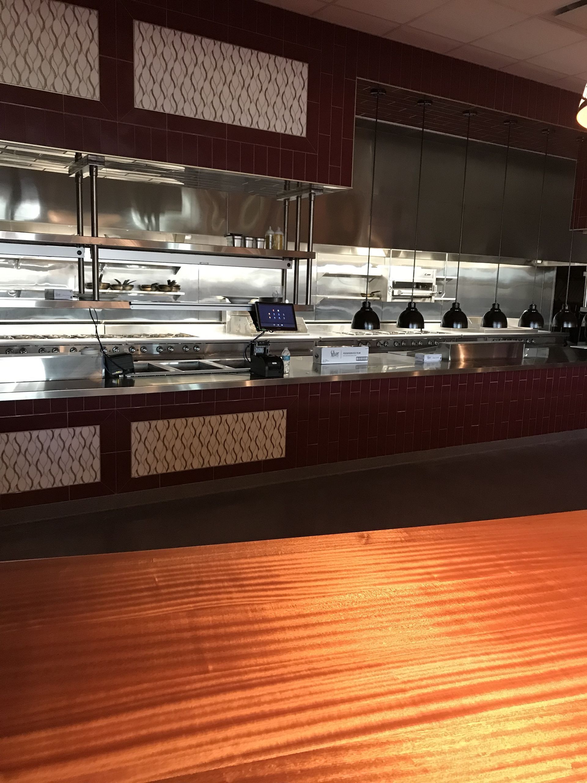 A wooden counter leads to a restaurant kitchen line featuring metallic surfaces, dark tiled walls, and patterned panels.