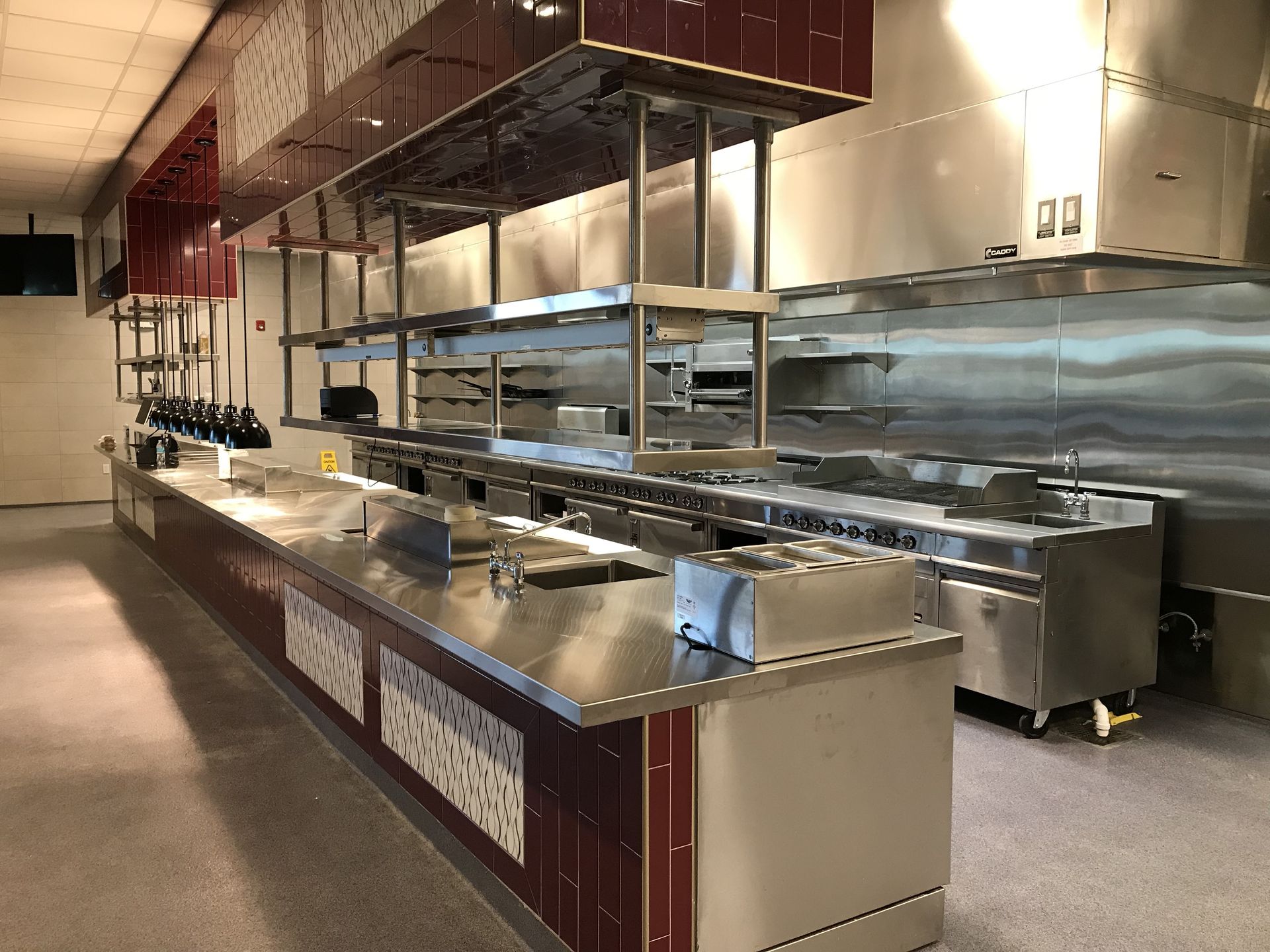 A long, commercial stainless steel kitchen counter and serving area with dark red tile accents and overhead shelving.