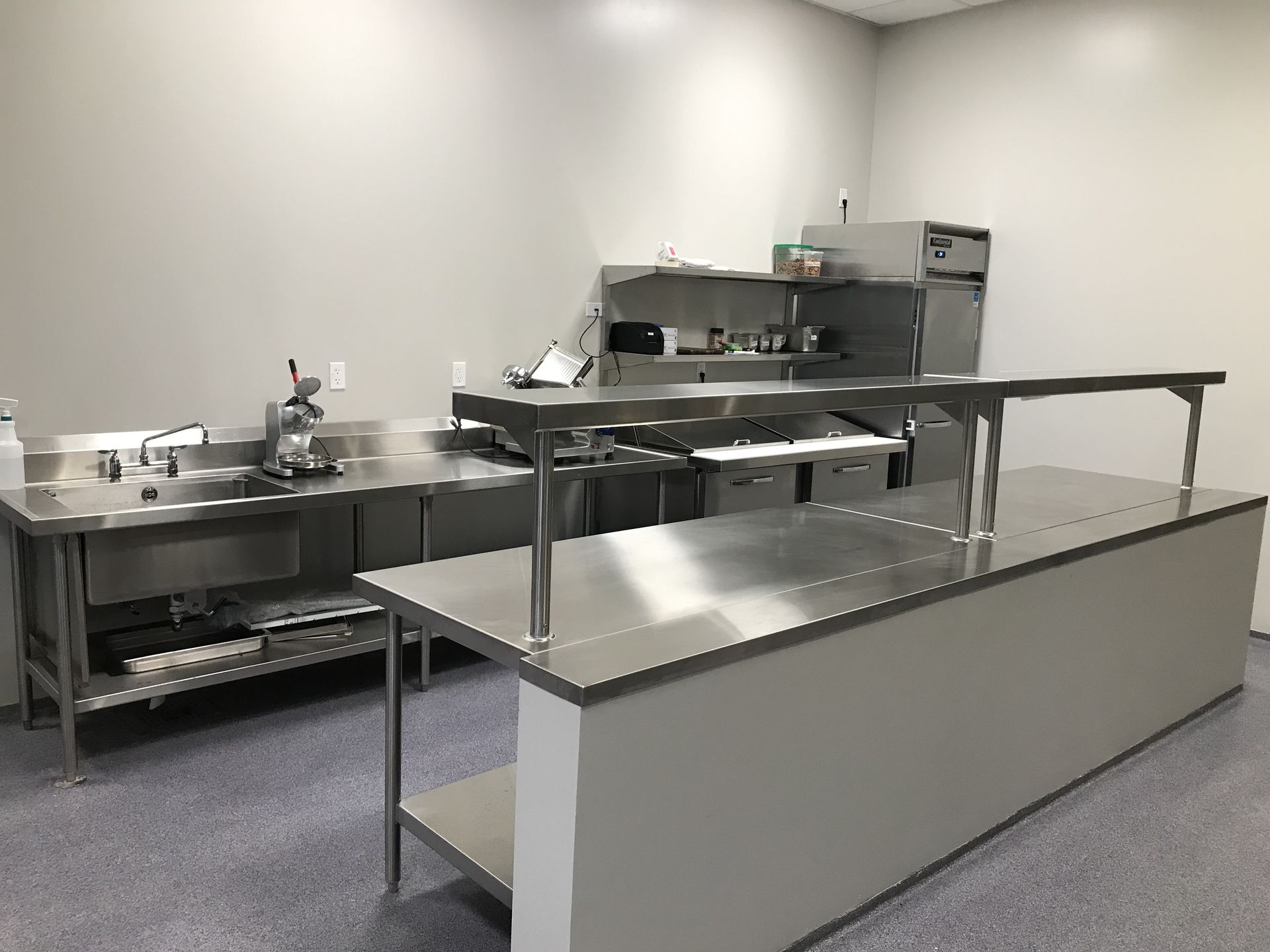 A commercial kitchen area featuring stainless steel prep tables, a sink, and cooking equipment on a speckled floor.