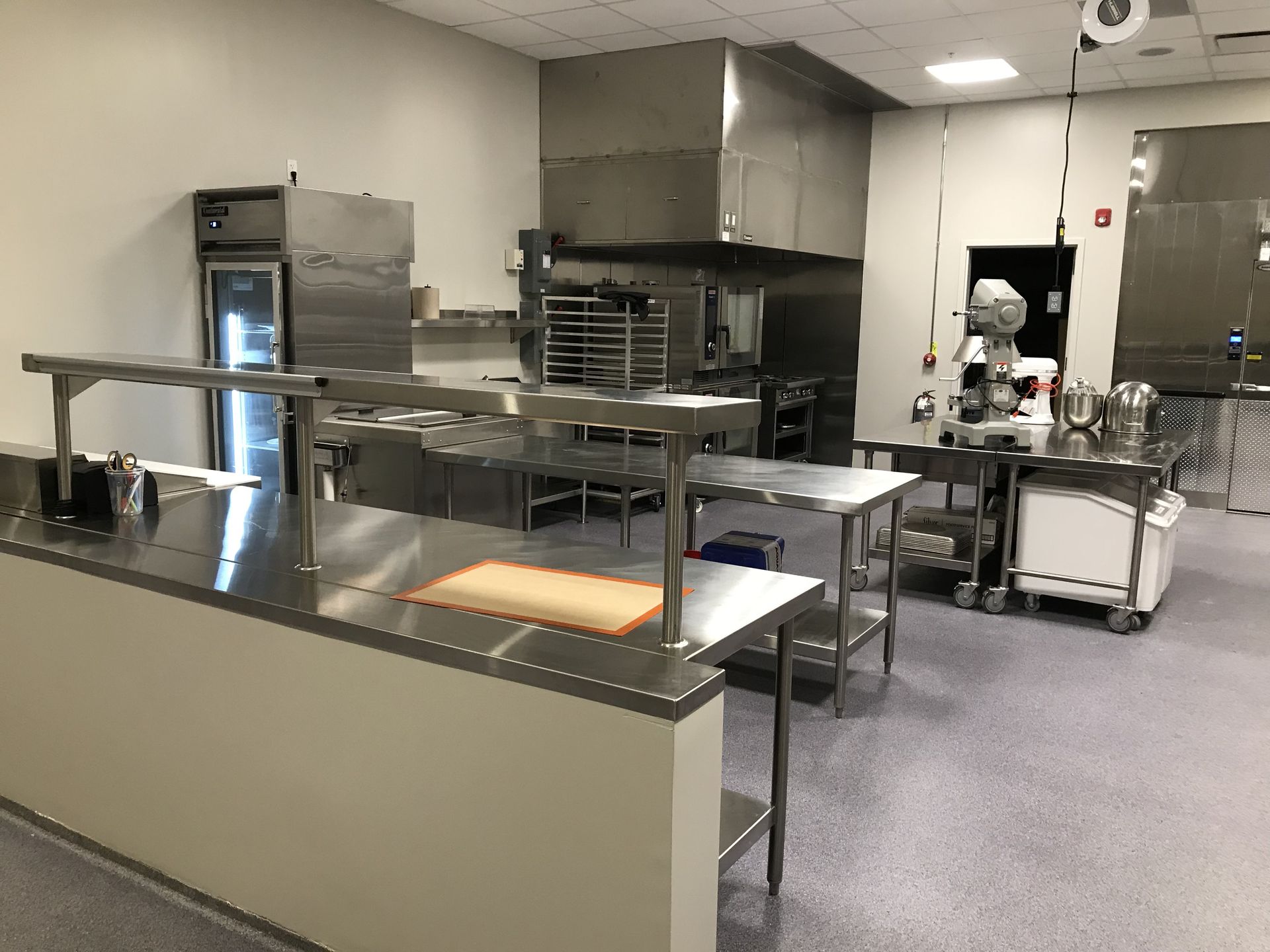 A clean, commercial kitchen with stainless steel workstations, a walk-in cooler, a large mixer, and grey speckled floors.