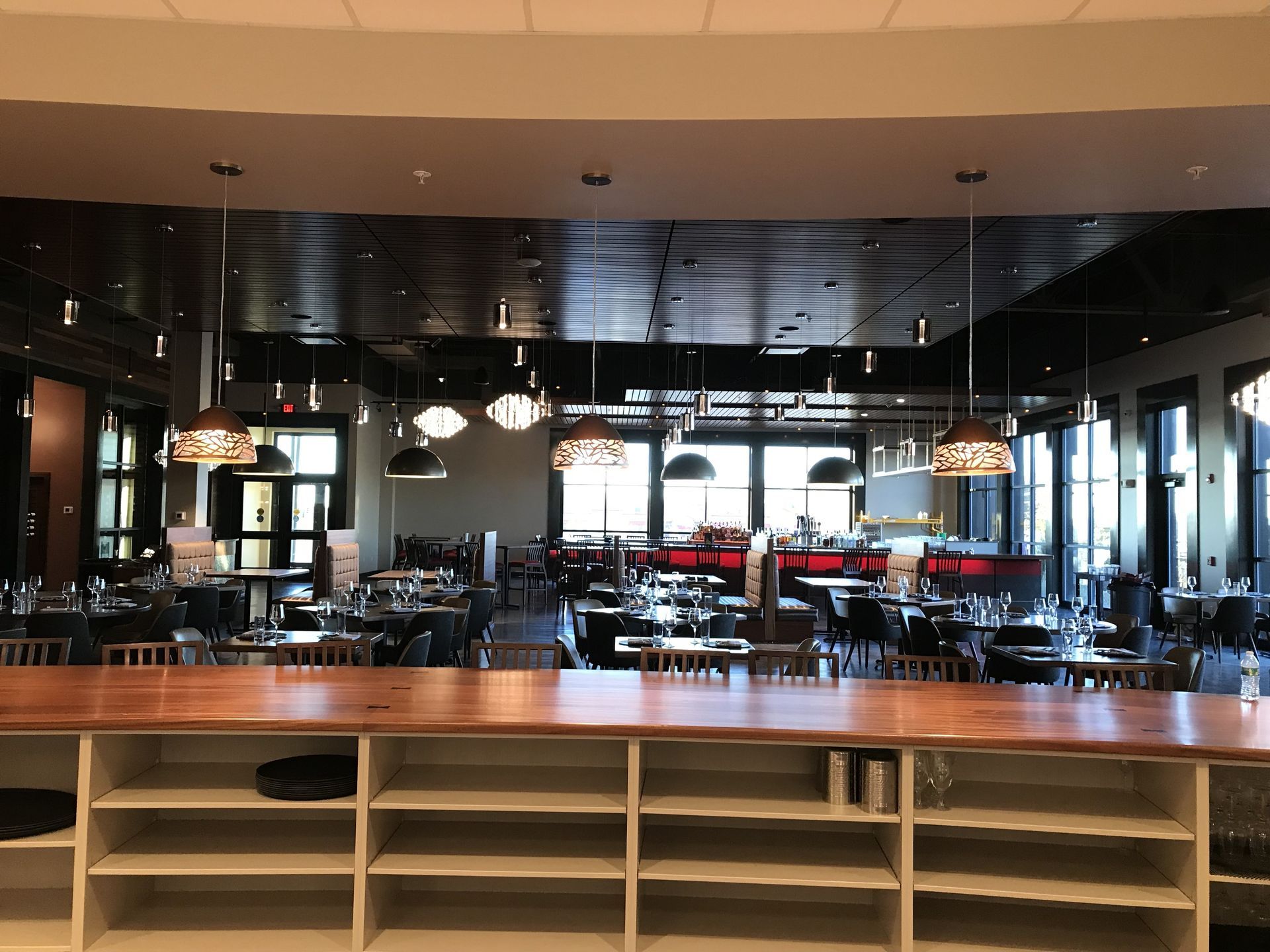 A long, wooden bar counter with white shelving faces a dining room with pendant lights, tables, and a dark ceiling.