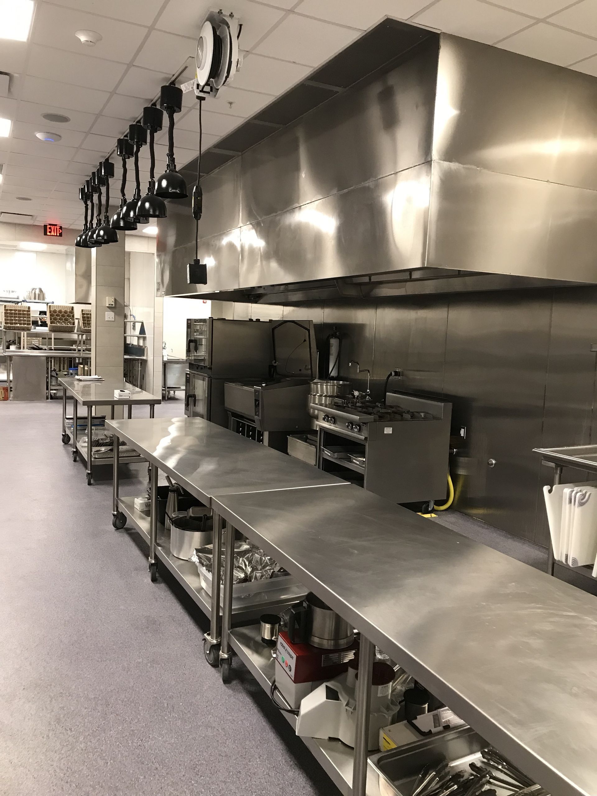 A clean, commercial kitchen with long stainless steel prep tables, cooking equipment under a large hood, and hanging lights.
