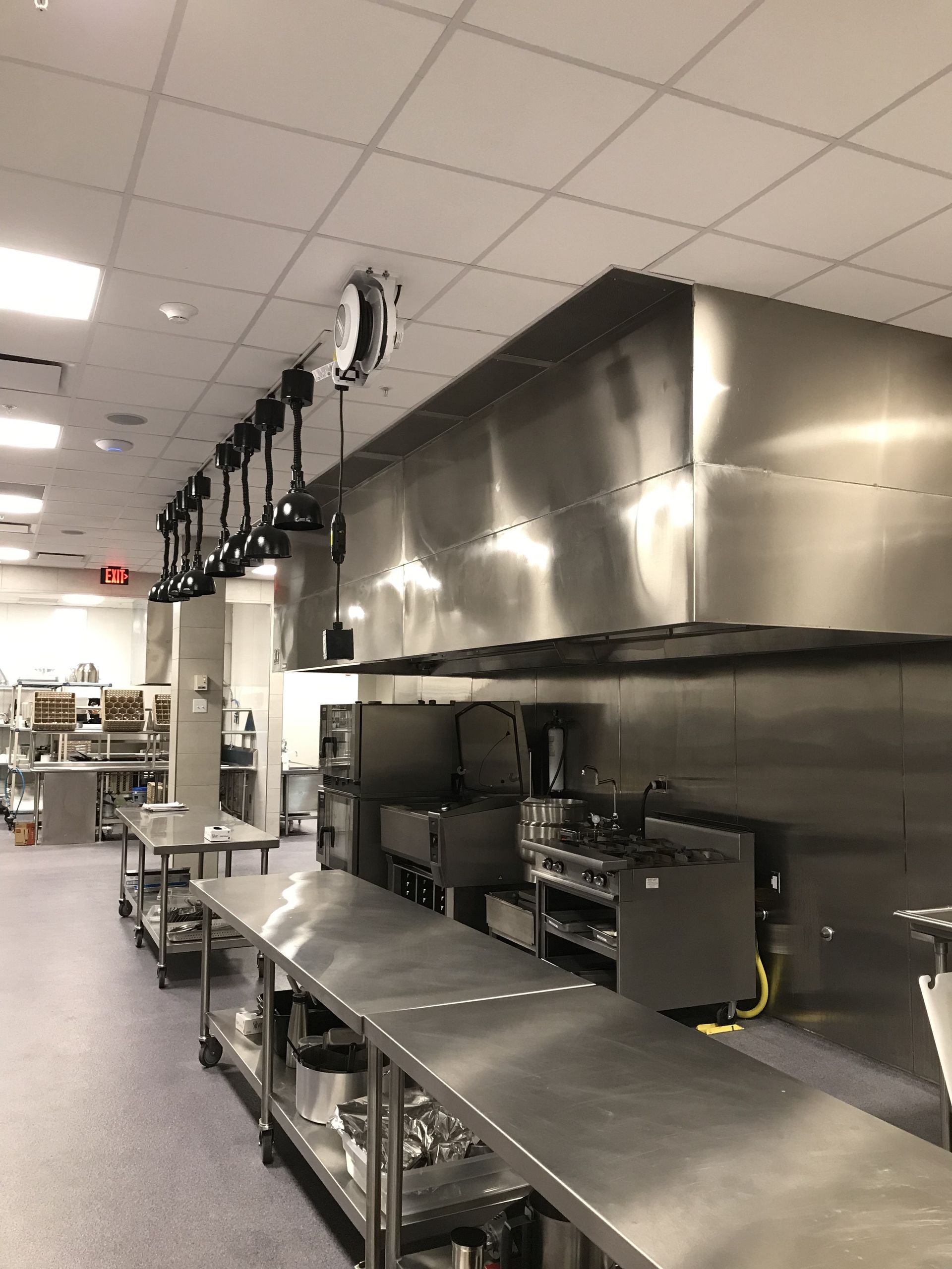 A commercial kitchen with stainless steel tables, a large overhead exhaust hood, and a row of hanging pendant lights.