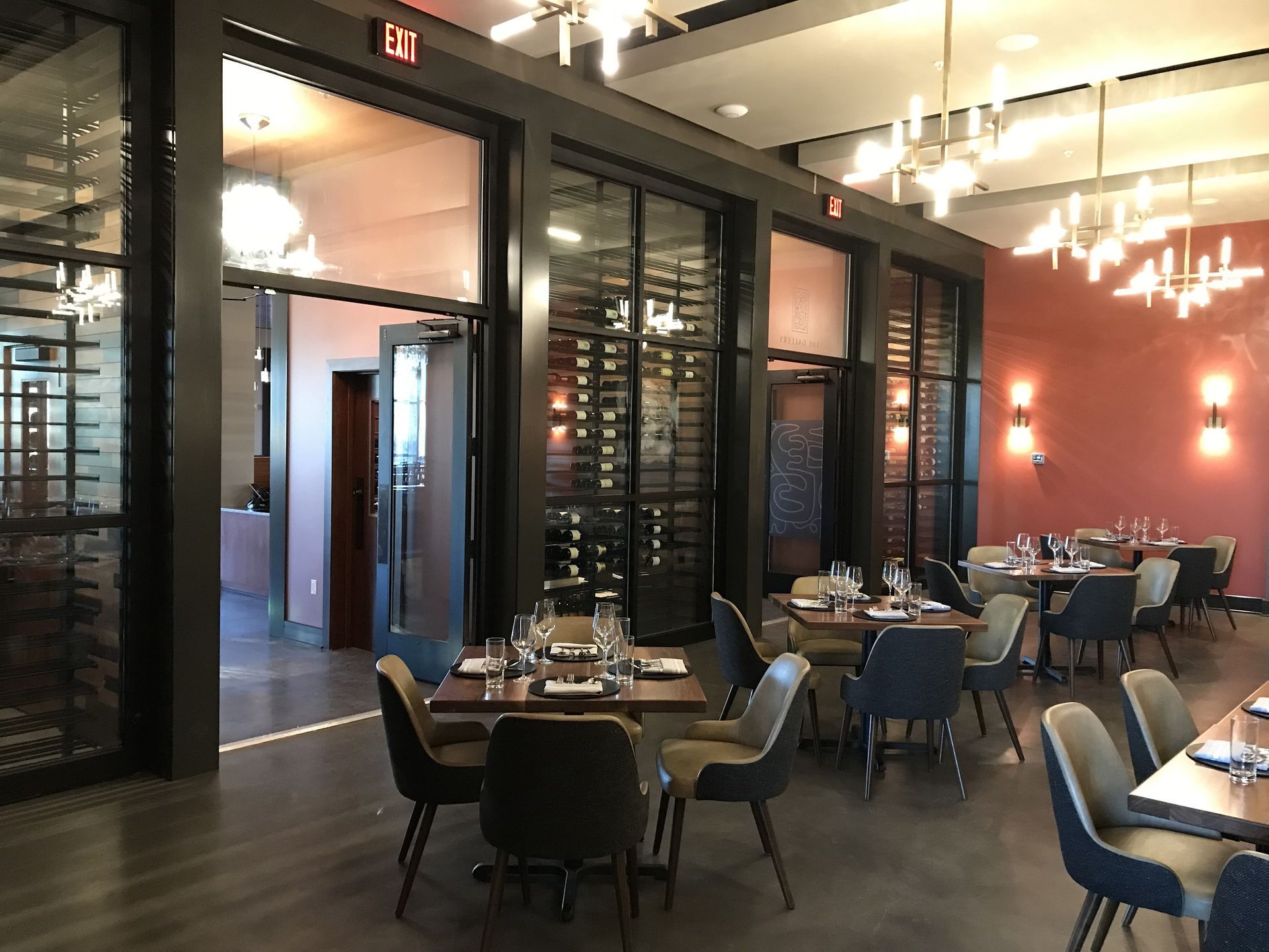 Modern dining area with dark wood tables, grey chairs, glass-enclosed wine storage, and warm lighting against peach walls.