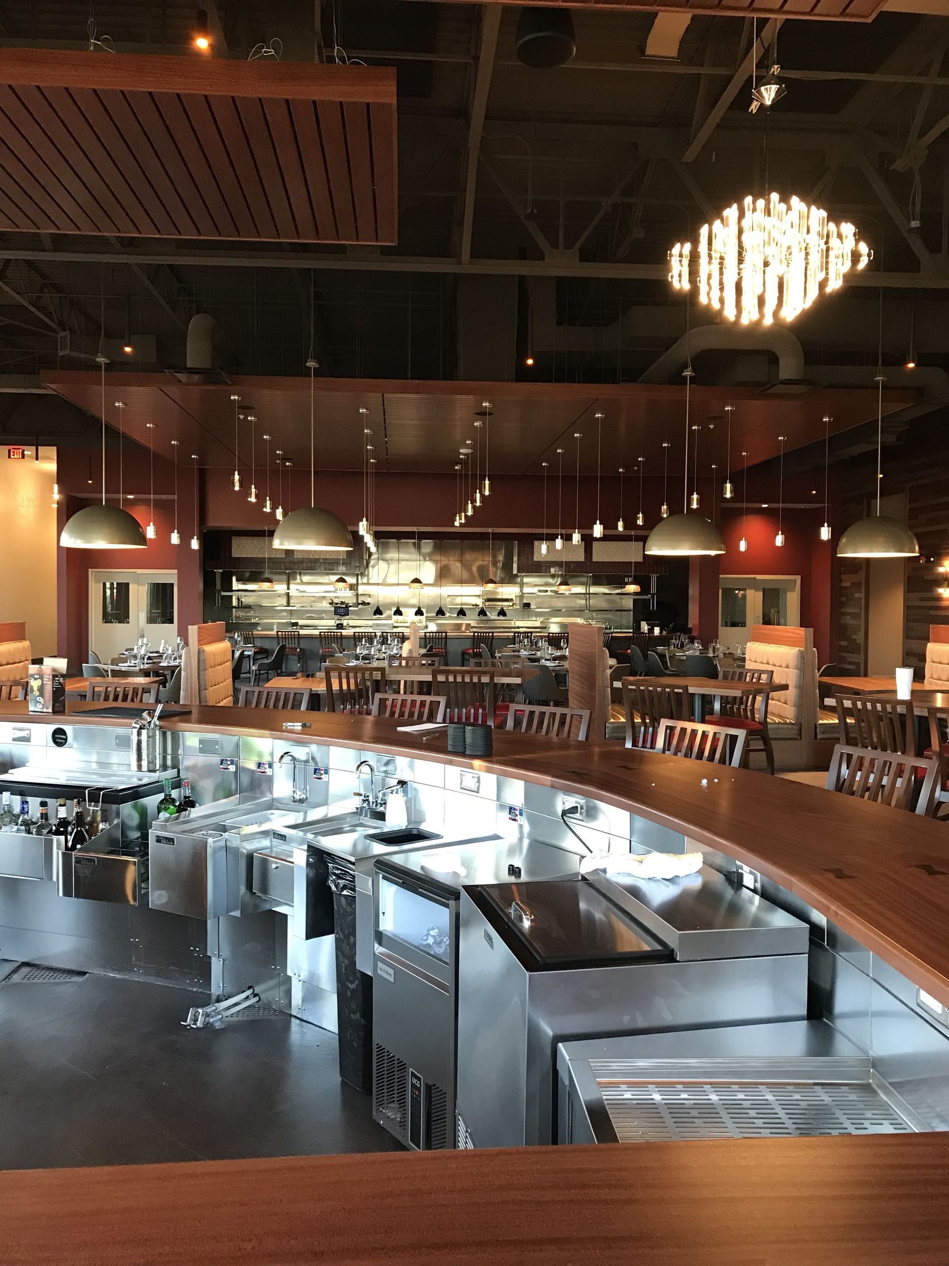 A modern restaurant interior featuring a polished wood bar counter, stainless steel prep equipment, and warm lighting.