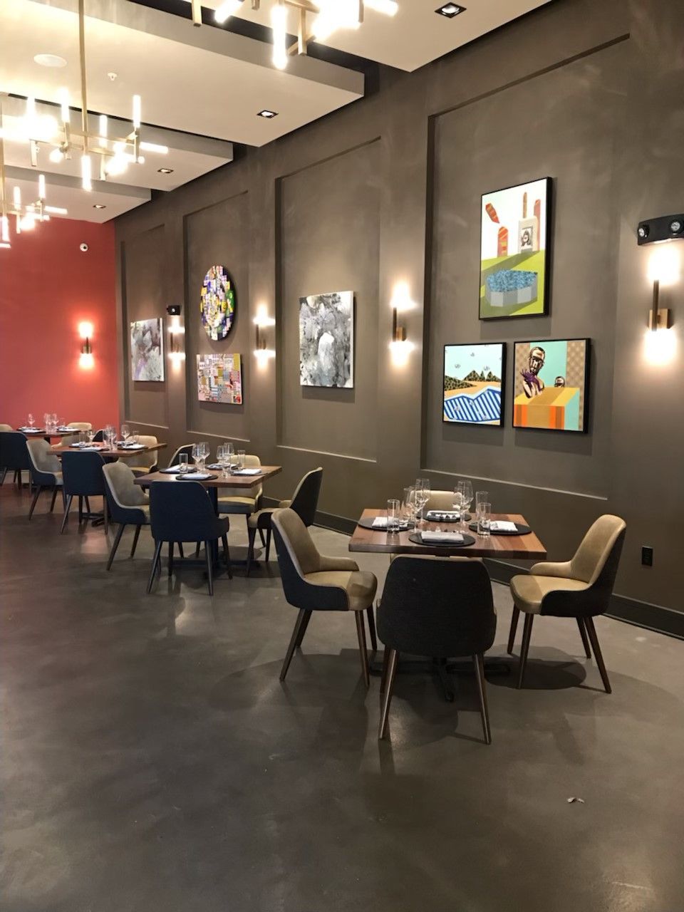 Modern restaurant dining room with dark walls, small tables, chairs, and abstract art displayed under wall sconces.