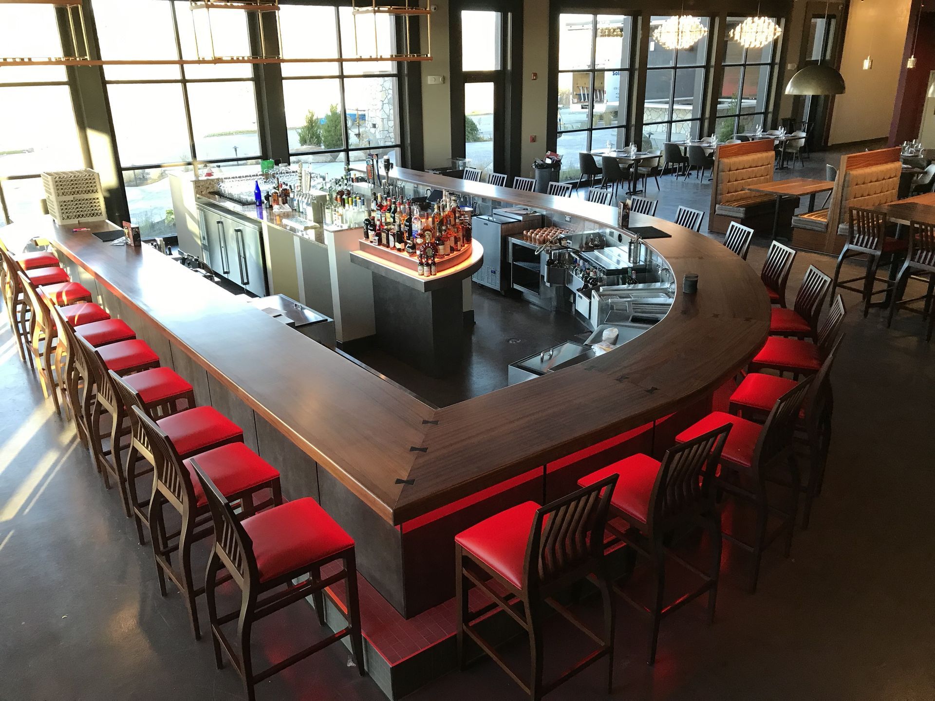 A U-shaped wooden bar with red bar stools in a restaurant setting with large windows.