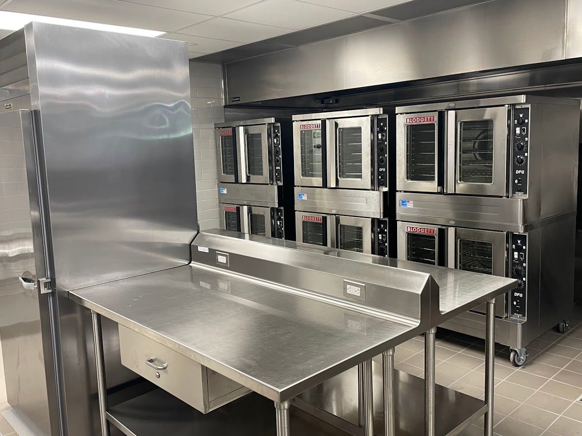 A commercial kitchen featuring stainless steel work tables in the foreground and a row of stacked industrial ovens behind.