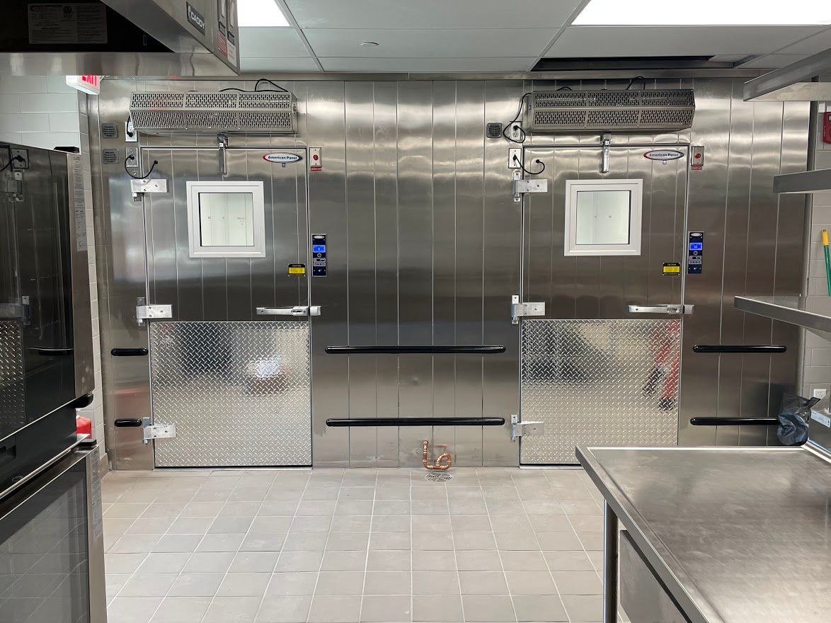 A commercial kitchen interior featuring two large, stainless steel walk-in cooler doors with small windows and metal plating.