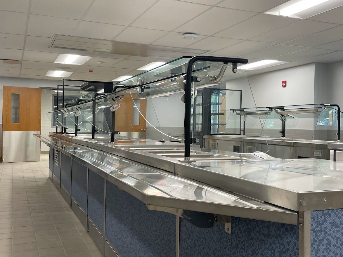 An empty cafeteria serving line with stainless steel food stations, sneeze guards, and blue patterned panels.