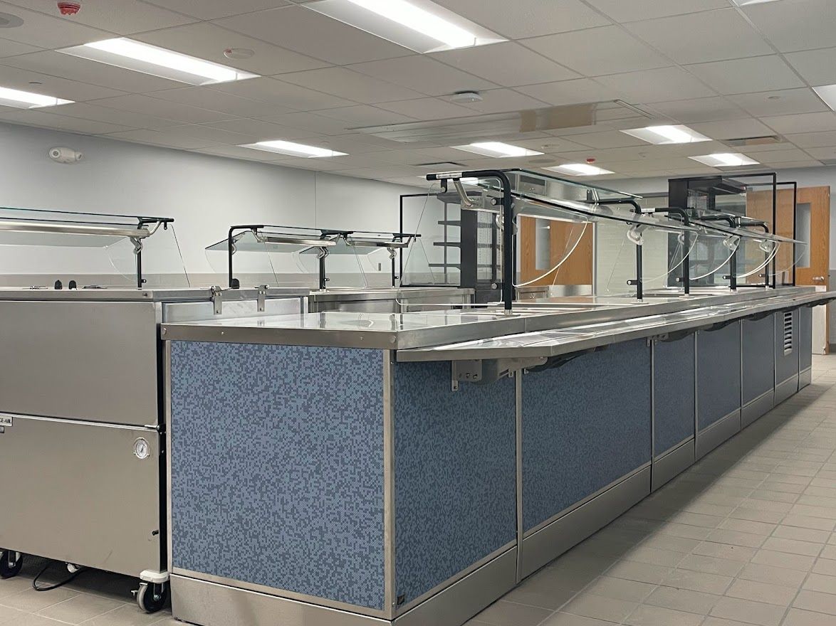 A long stainless steel cafeteria serving counter with blue speckled panels in a brightly lit, commercial kitchen space.