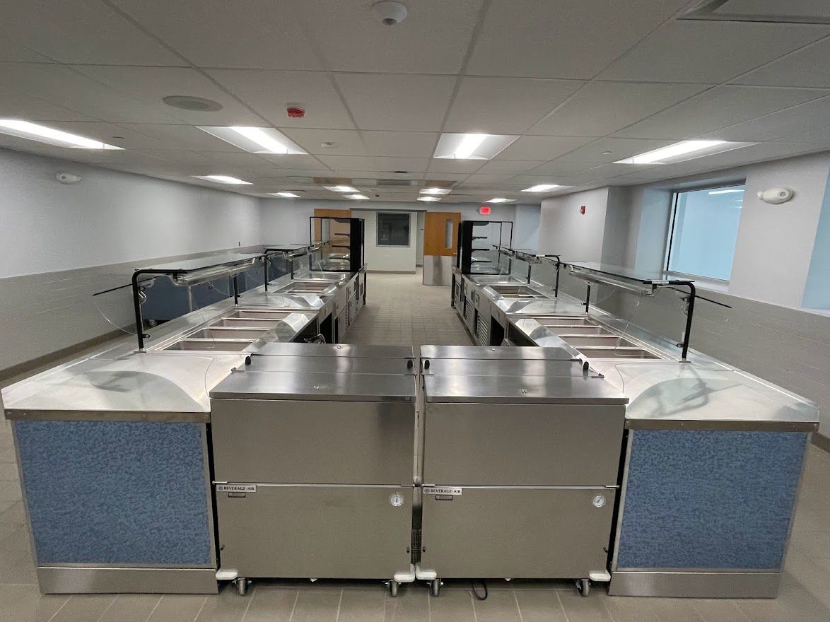 A commercial stainless steel serving counter in an empty, brightly lit cafeteria with light blue textured paneling.