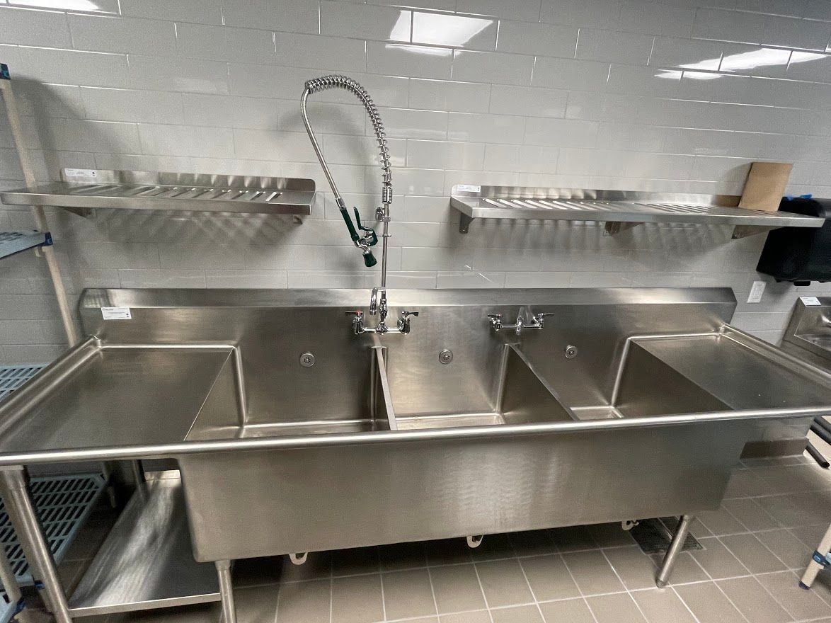 A large, three-compartment stainless steel industrial sink with a spray faucet mounted against a white tiled wall.