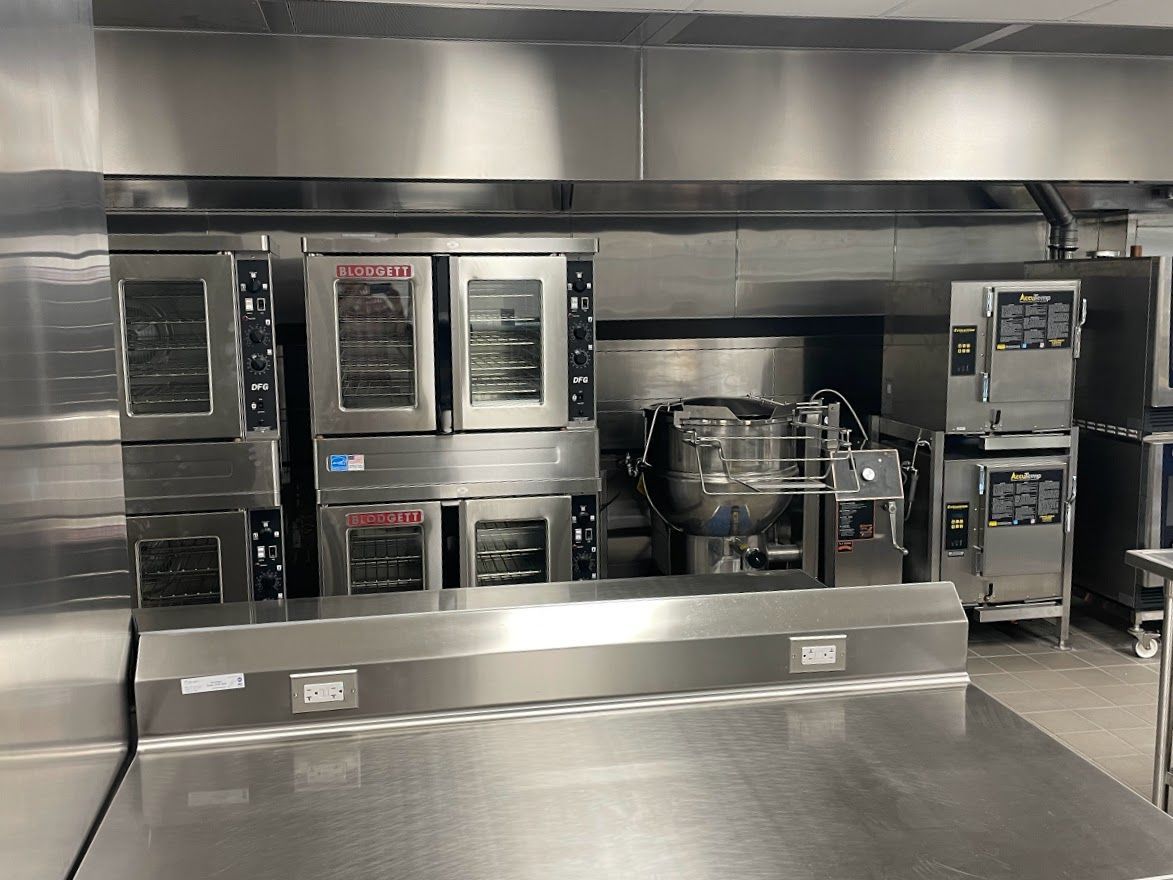 A professional stainless steel kitchen with stacked commercial convection ovens, a tilting skillet, and a steam kettle.