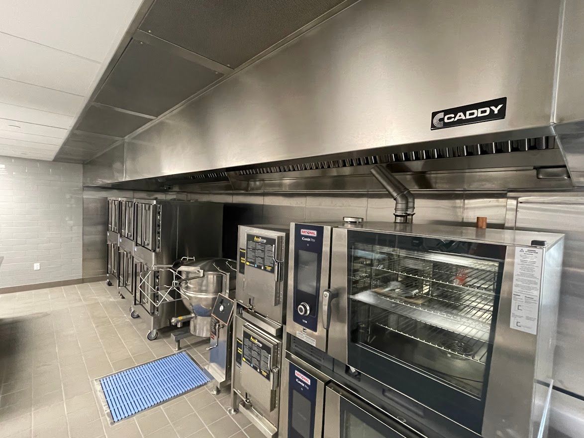 A stainless steel commercial kitchen featuring rows of professional ovens and a Caddy ventilation hood above.