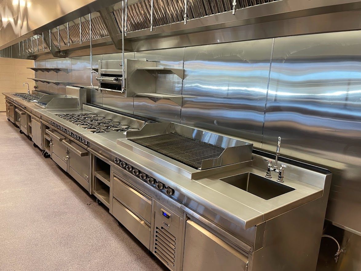 A stainless steel commercial kitchen station with a stove, grill, prep surface, and sink under a ventilation hood.