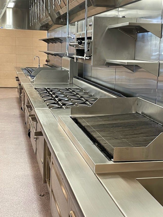 A professional stainless-steel commercial kitchen prep line featuring a grill, gas burners, and a sink.