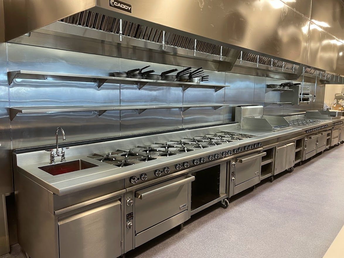 A stainless steel commercial kitchen line with a sink, burners, ovens, and overhead ventilation hood.