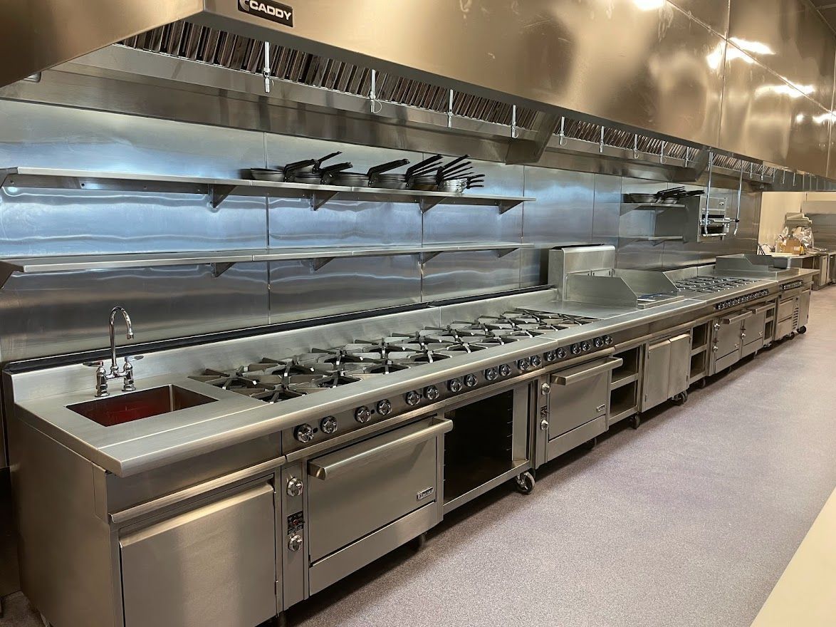 A clean, stainless steel commercial kitchen line with a built-in sink, stove burners, and overhead ventilation.