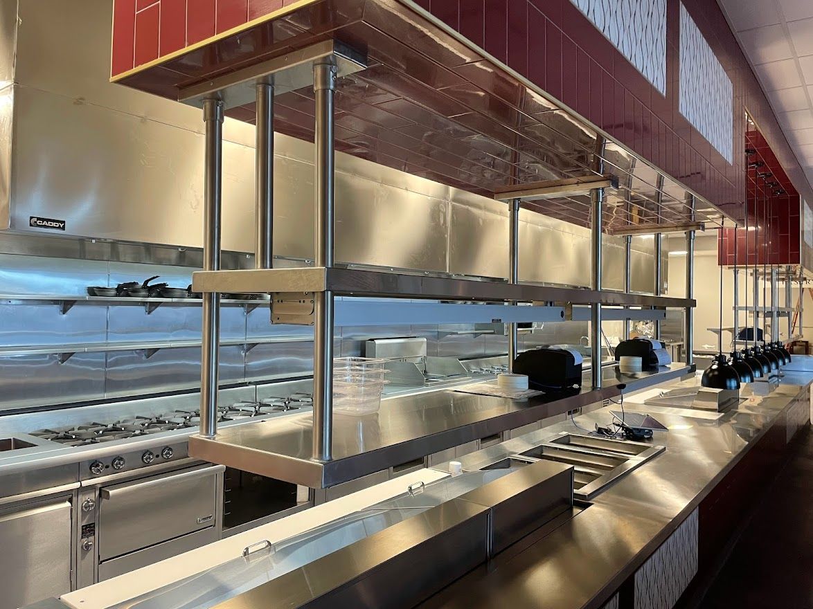 A commercial kitchen pass with stainless steel counters and shelving under a red-tiled ventilation hood.
