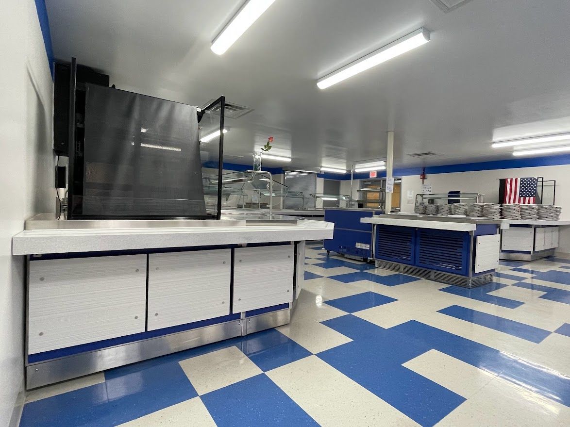 An empty school cafeteria with stainless steel serving counters, white cabinets, and a blue and white checkered floor.