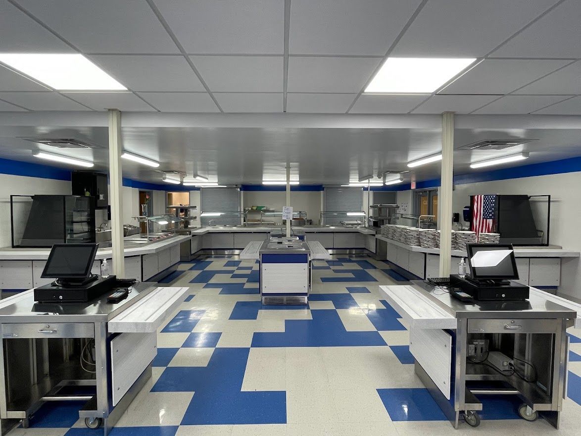 An empty school cafeteria with stainless steel food service counters, black cash registers, and a blue and white floor.
