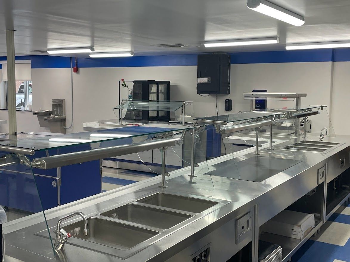 A stainless steel cafeteria serving line with a three-basin sink, glass sneeze guards, and blue-and-white tiled walls.