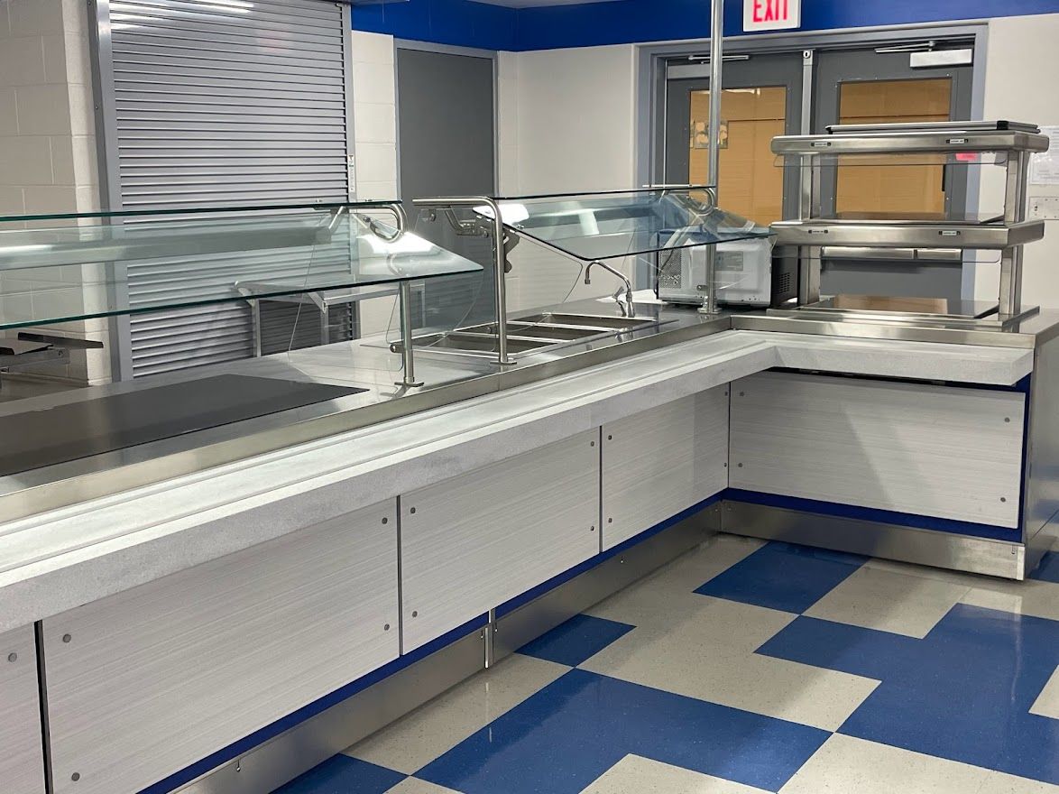 A stainless steel cafeteria serving counter with a glass sneeze guard, set against a blue and white tiled floor.