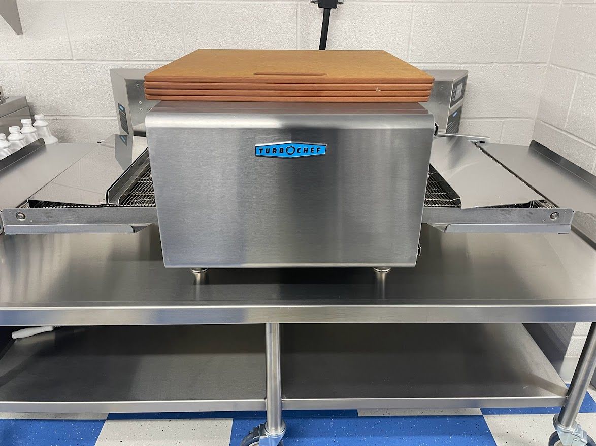 A commercial stainless steel conveyor toaster sits on a metal table in a kitchen.