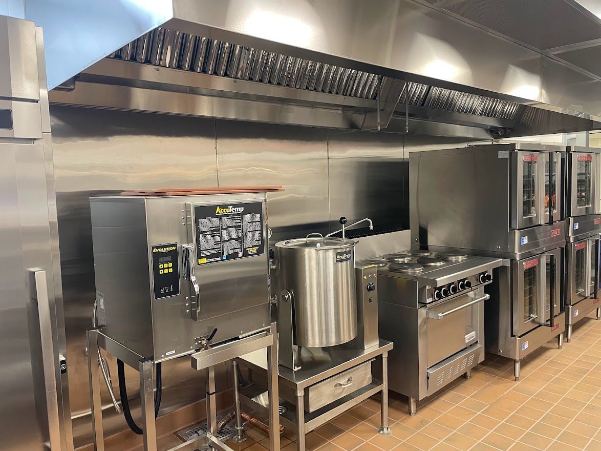 Stainless steel commercial kitchen equipment, including a steam oven, tilt skillet, and ranges, under a ventilation hood.