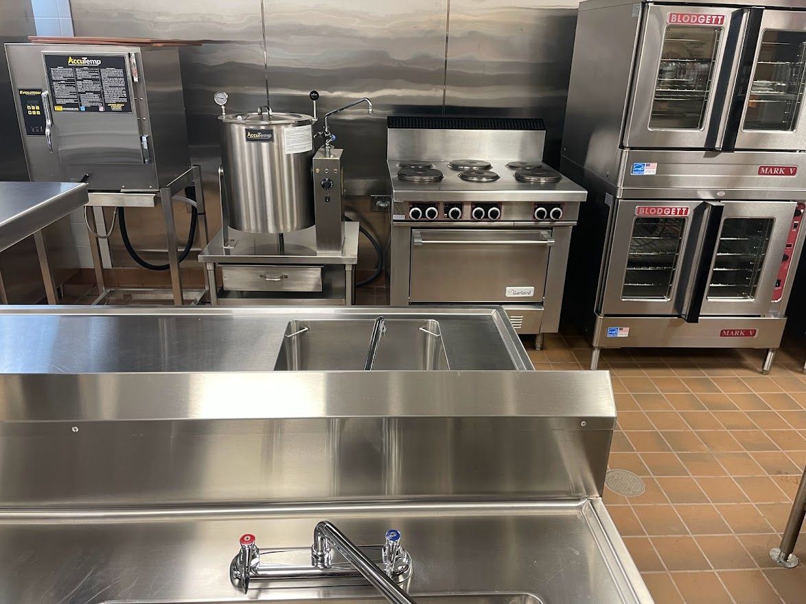 Commercial kitchen with stainless steel appliances, a sink, a stove, and stacked ovens.