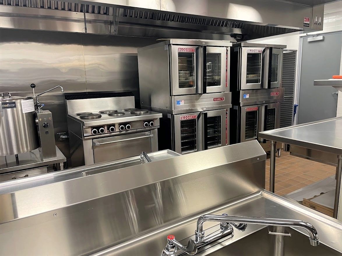 A commercial kitchen featuring stainless steel prep tables, a sink, a gas range, and stacked convection ovens.