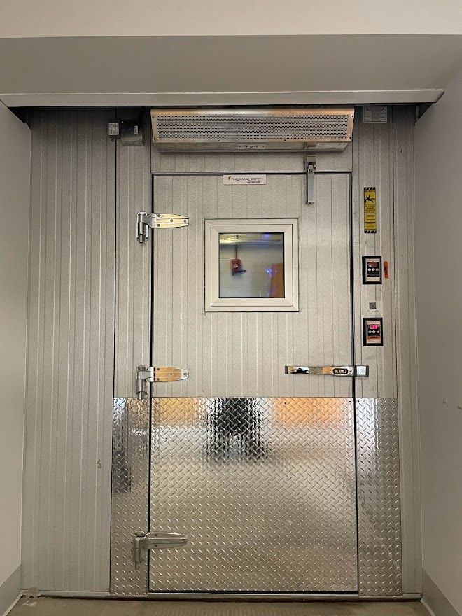 A walk-in freezer door with a small window, diamond-plate kick plate, and silver metal finish, set in a plain wall.