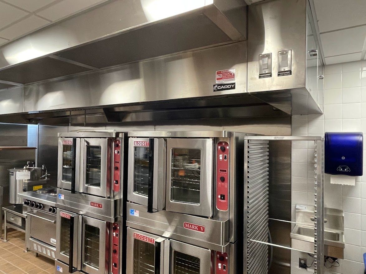 Commercial kitchen with stainless steel convection ovens under a large exhaust hood and a wall-mounted towel dispenser.