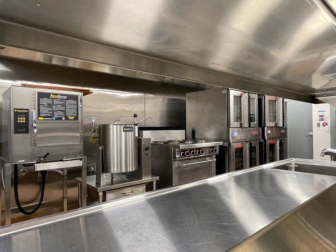 A stainless steel commercial kitchen featuring a steamer, tilt skillet, range, and stacked convection ovens under a hood.
